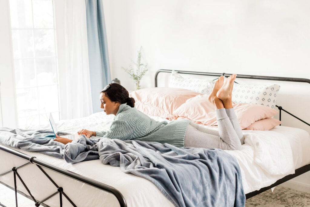 An image of a black woman lying on her bed with a laptop on a blog post about support for modern motherhood from a Boston mom therapist.